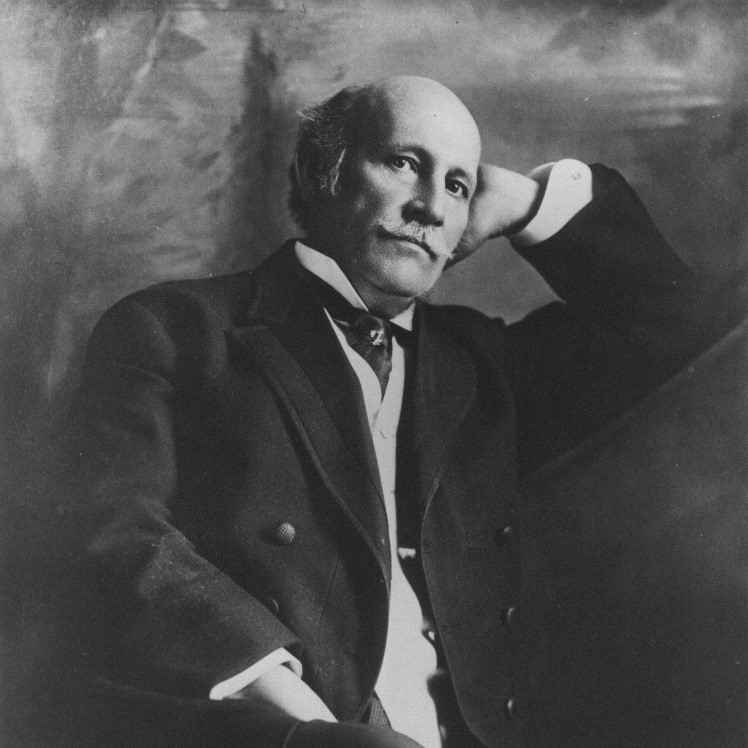 Black-and-white portrait of an older man with a receding hairline and mustache, wearing a suit, resting his head on his right hand.