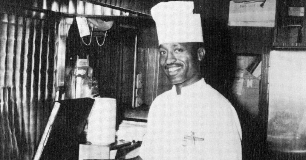 | Tennessee Wesleyan University Smiling Black chef in white uniform and tall hat stands in a busy kitchen beside a counter.