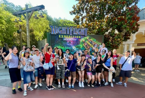 Group of smiling people posing together outdoors in front of a colorful sign that reads 'Nightlight Expedition' with a ride poster behind them.