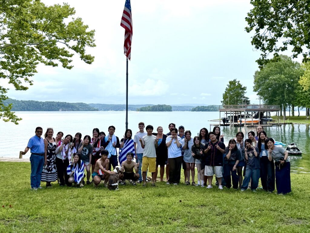 | Tennessee Wesleyan University Group photo of many people standing on grass by a lake, with an American flag on a tall pole in the background.