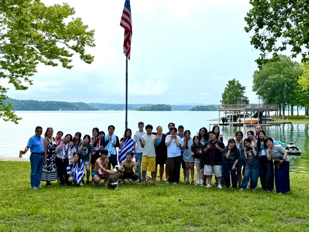 Group photo of many people standing on grass by a lake, with an American flag on a tall pole in the background.