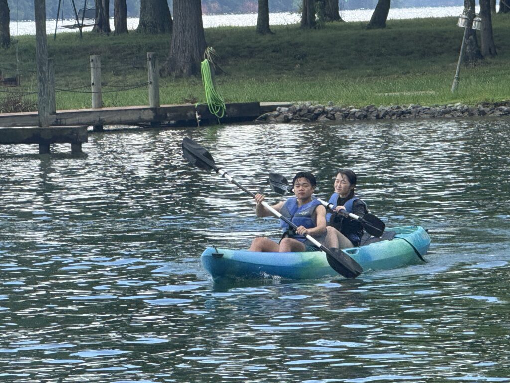 Two people with life jackets Kayaking in a blue tandem kayak near a wooden dock on a lake, with a neon green rope hanging from a post and trees in the background.