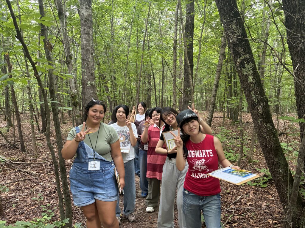 | Tennessee Wesleyan University Group of smiling people posing in a forest, waving and making peace signs among trees and fallen leaves.
