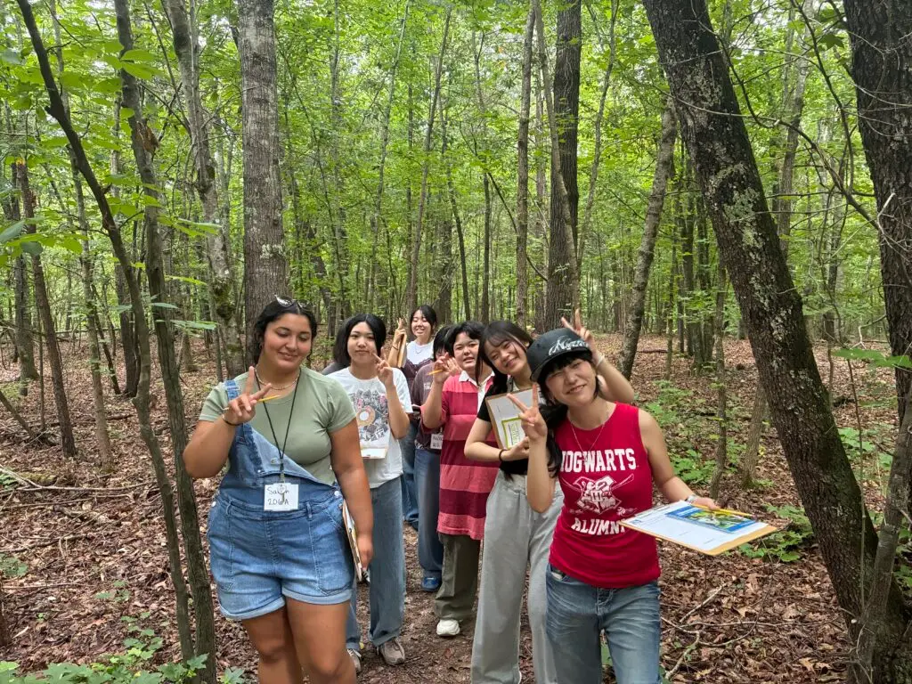 Group of smiling people posing in a forest, waving and making peace signs among trees and fallen leaves.