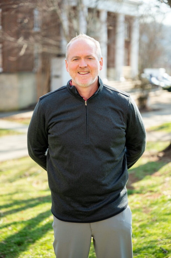 | Tennessee Wesleyan University Smiling man with short gray hair wearing a dark quarter-zip sweater outdoors on a sunny day.