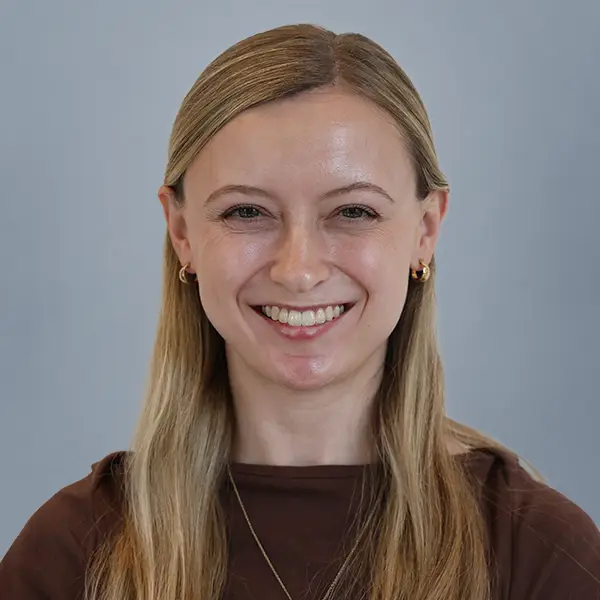 Portrait of a smiling woman with long blonde hair wearing a brown top against a blue-gray background, looking at the camera.