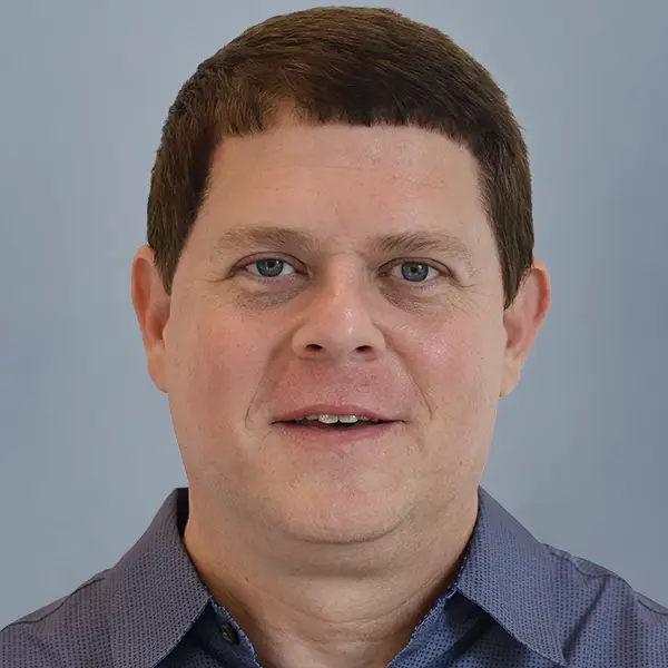Head-and-shoulders portrait of a man with short brown hair wearing a blue collared shirt, neutral expression, gray background.