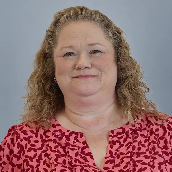 Smiling woman with shoulder-length curly blond hair wearing a pink patterned blouse against a gray backdrop.