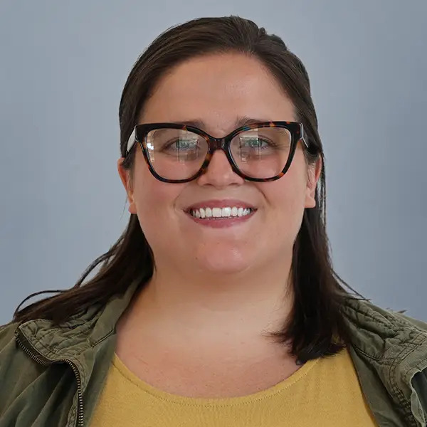 Portrait of a smiling woman with dark hair, wearing tortoiseshell glasses and a mustard-yellow top with an olive jacket, against a blue-gray background.