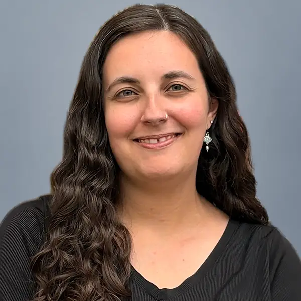 Portrait of a smiling woman with long dark hair and a nose ring, wearing a black top against a blue-gray background.