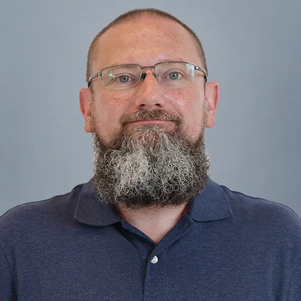 Portrait of a man with glasses and a gray beard wearing a navy polo against a gray backdrop.