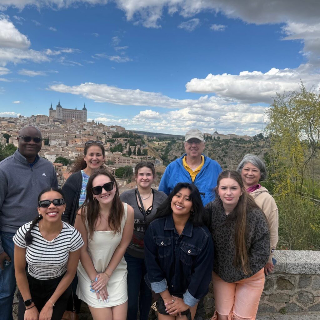 Group of smiling people posing for a photo with a historic cityscape and castle in the background on a sunny day