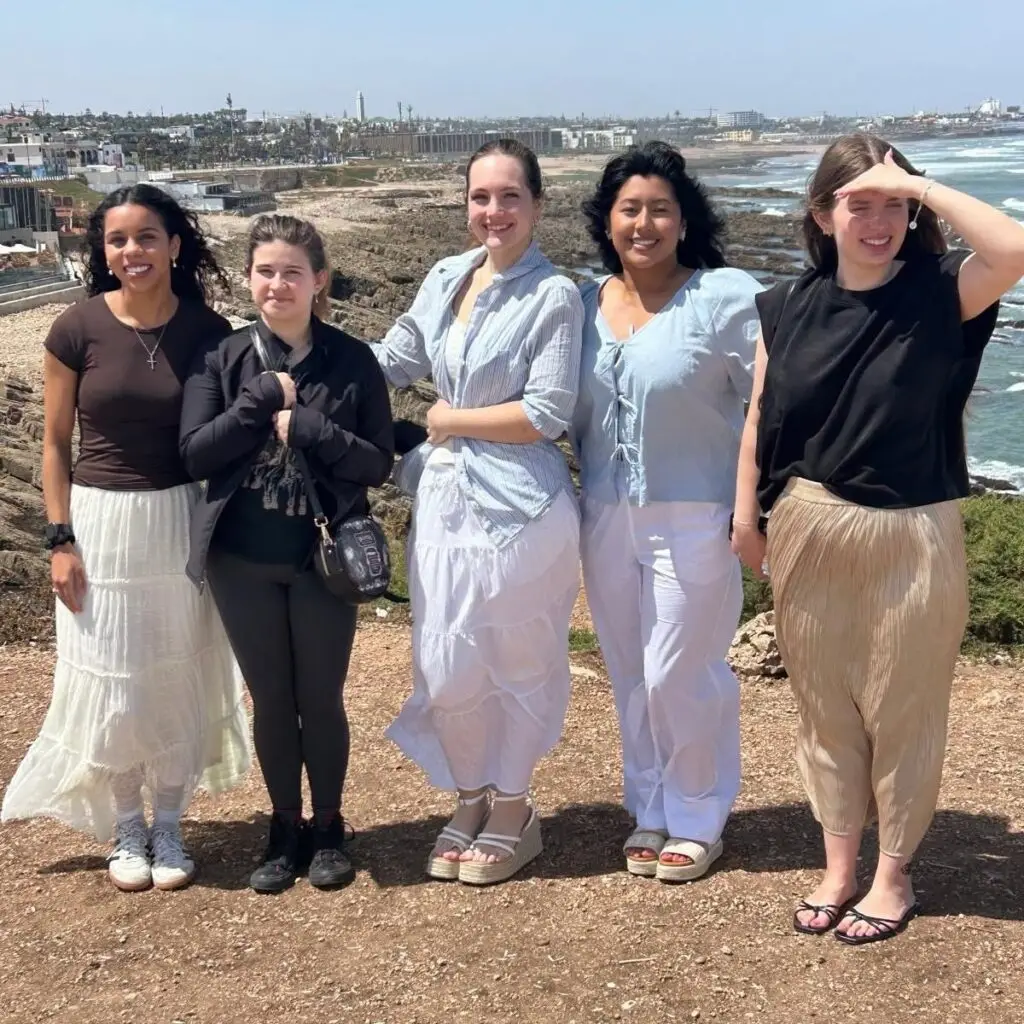 Five women stand on a rocky coastal overlook, smiling at the camera; they wear casual summer outfits near the ocean.