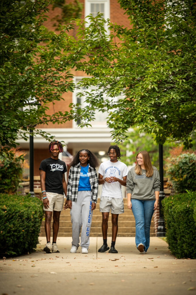 | Tennessee Wesleyan University Four students walking down a campus sidewalk framed by green trees with a brick building in the background.