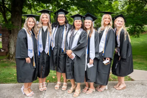 Seven graduates in black caps and gowns, outdoors, smiling and posing with diplomas and cords in a park setting.