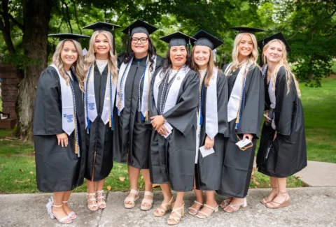 Seven graduates in black caps and gowns, outdoors, smiling and posing with diplomas and cords in a park setting.