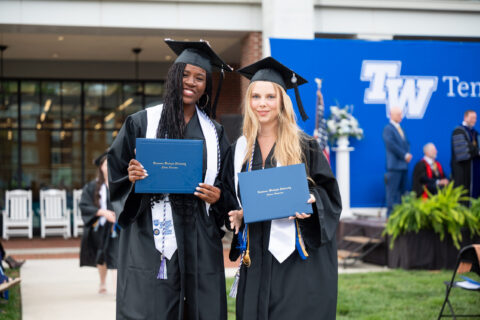 Two graduates in caps and gowns hold blue diploma folders and smile at an outdoor ceremony.