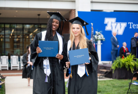 Two graduates in caps and gowns hold blue diploma folders and smile at an outdoor ceremony.