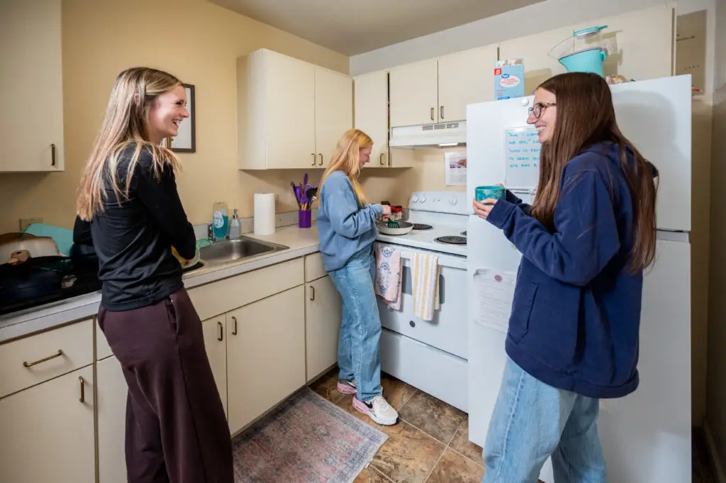 Three young women in a kitchen smiling and chatting: one at the sink, one at the stove, and another by the refrigerator.