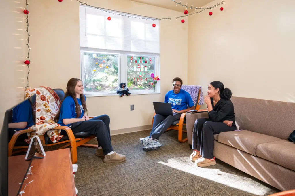 Three people sit in a cozy living room chatting, Christmas lights and a window with decorations above them.