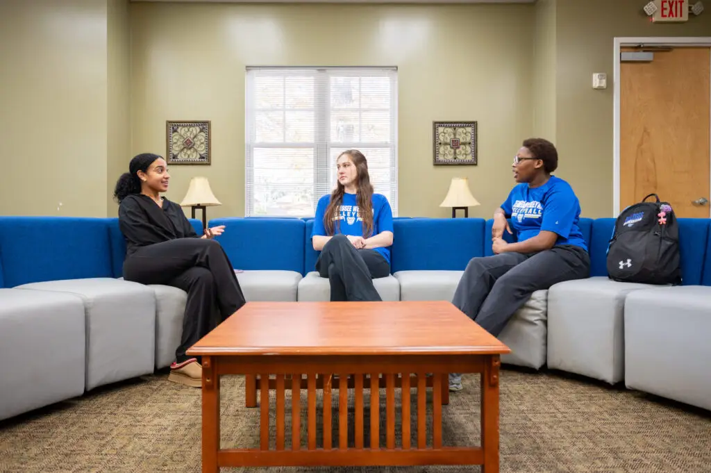 Three people sit on a blue sectional discussing around a wooden coffee table in a lounge area.