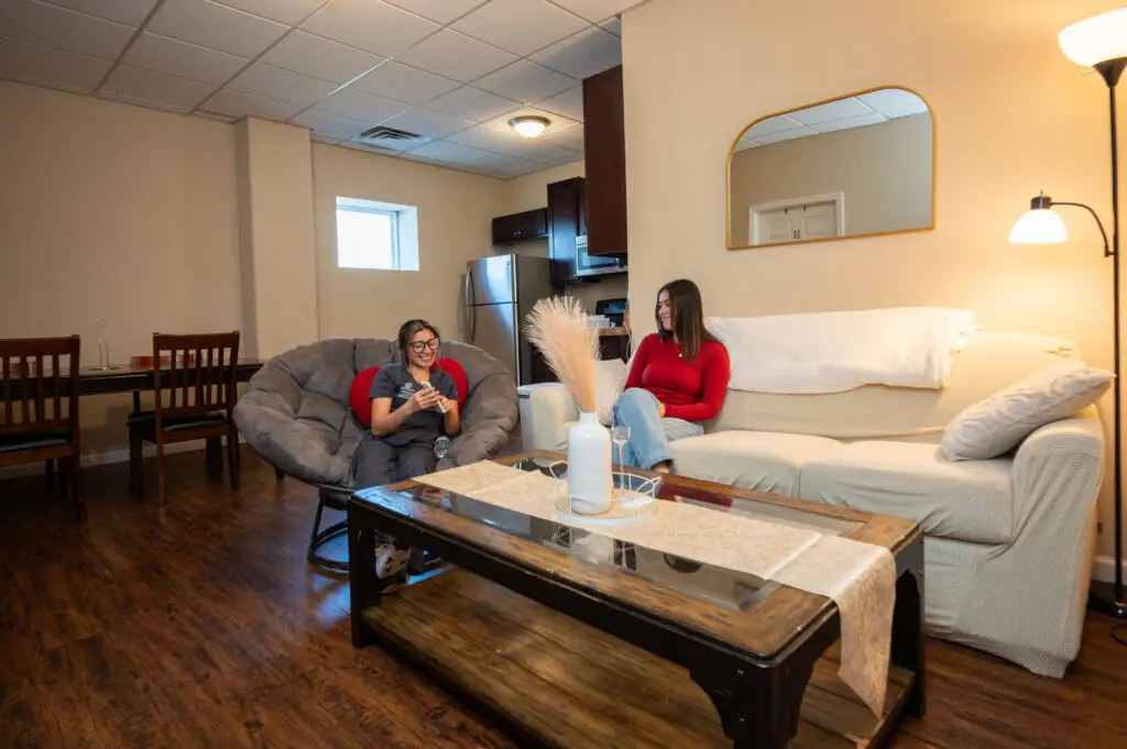 Two women sit in a cozy living room: one on a gray papasan chair reading, the other on a cream sofa chatting; a kitchen is in the back.