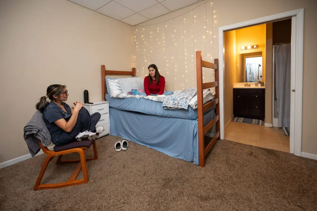 Dorm room with two wooden bunk beds, fairy lights on the wall, a girl in a red shirt sits on the lower bed while another person sits nearby in a chair.