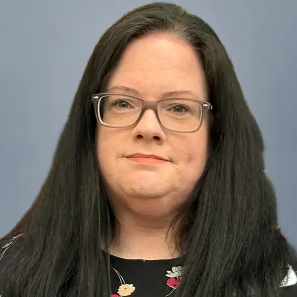 Portrait of a woman with long dark hair and rectangular glasses, wearing a black floral top, against a blue-gray background.