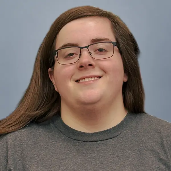 Smiling person with long dark hair and rectangular glasses, wearing a gray crewneck shirt, against a blue-gray background.