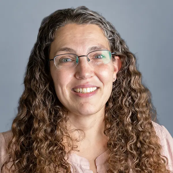 Smiling woman with long, curly hair and glasses, wearing a light pink top, against a gray background.