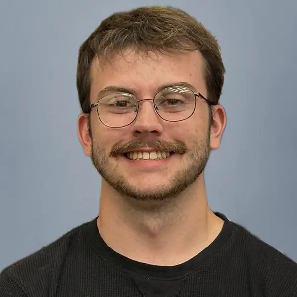 Smiling young man with short brown hair and round glasses, wearing a black shirt against a light-blue background.