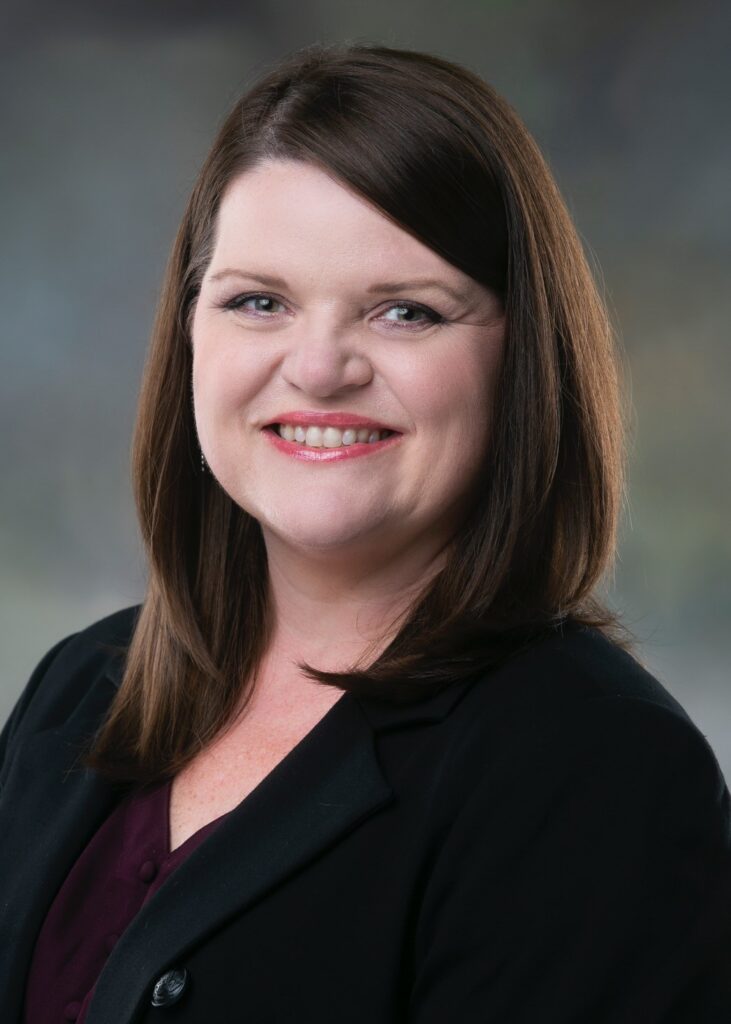 Smiling woman wearing a black blazer; shoulder-length brown hair, professional headshot.