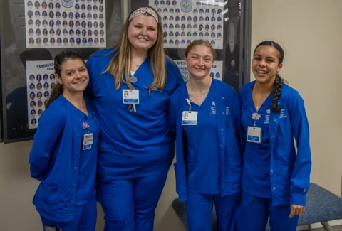 Four women in blue scrubs stand together in a hallway, smiling, with badge IDs; posters on the wall behind them.