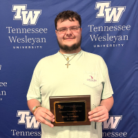 Man in a light green shirt holding a wooden plaque in front of a Tennessee Wesleyan backdrop, wearing a gold cross necklace and beaded bracelet, smiling for the photo.